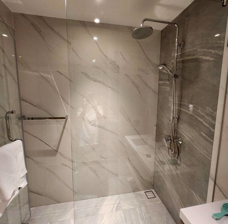 Glass-walled walk-in shower with large white and gray marble tiles, a chrome rainfall showerhead, handheld fixture, and towel bar in a modern bathroom.