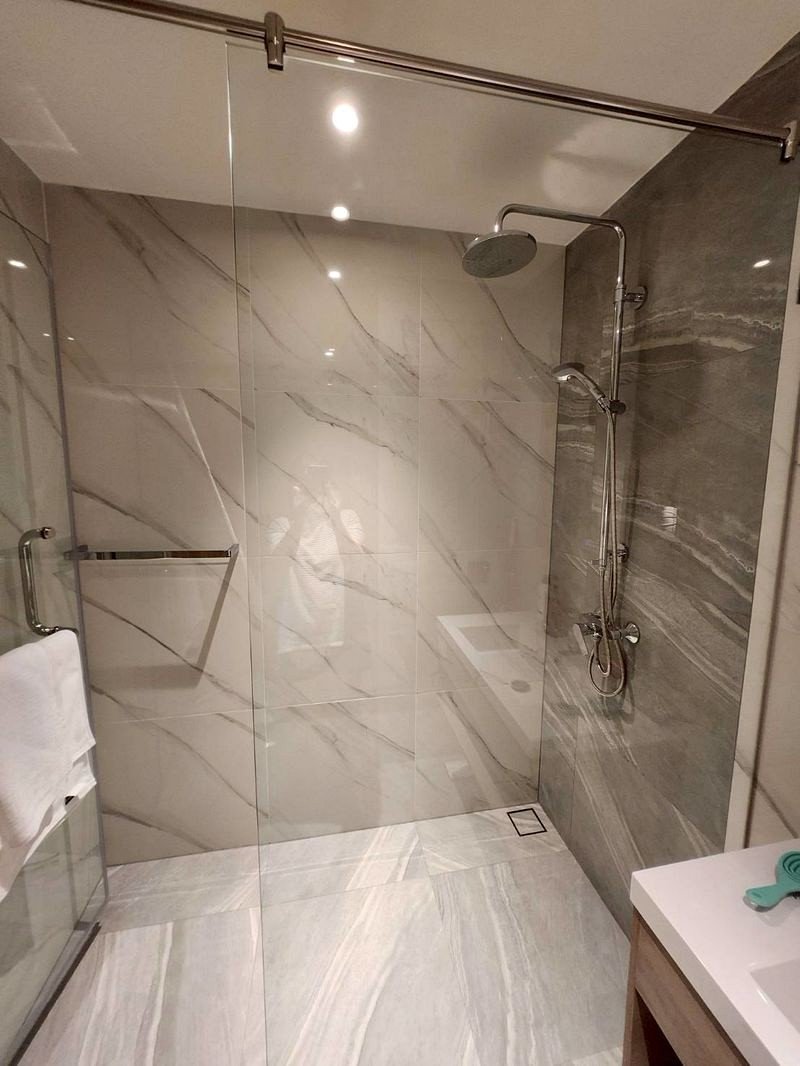 Glass-walled walk-in shower with large white and gray marble tiles, a chrome rainfall showerhead, handheld fixture, and towel bar in a modern bathroom.