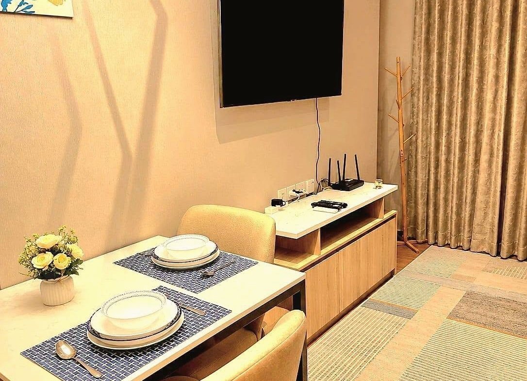 Dining area with two beige upholstered chairs and a white table set with plates, a vase of white flowers, and blue placemats; wall-mounted TV nearby