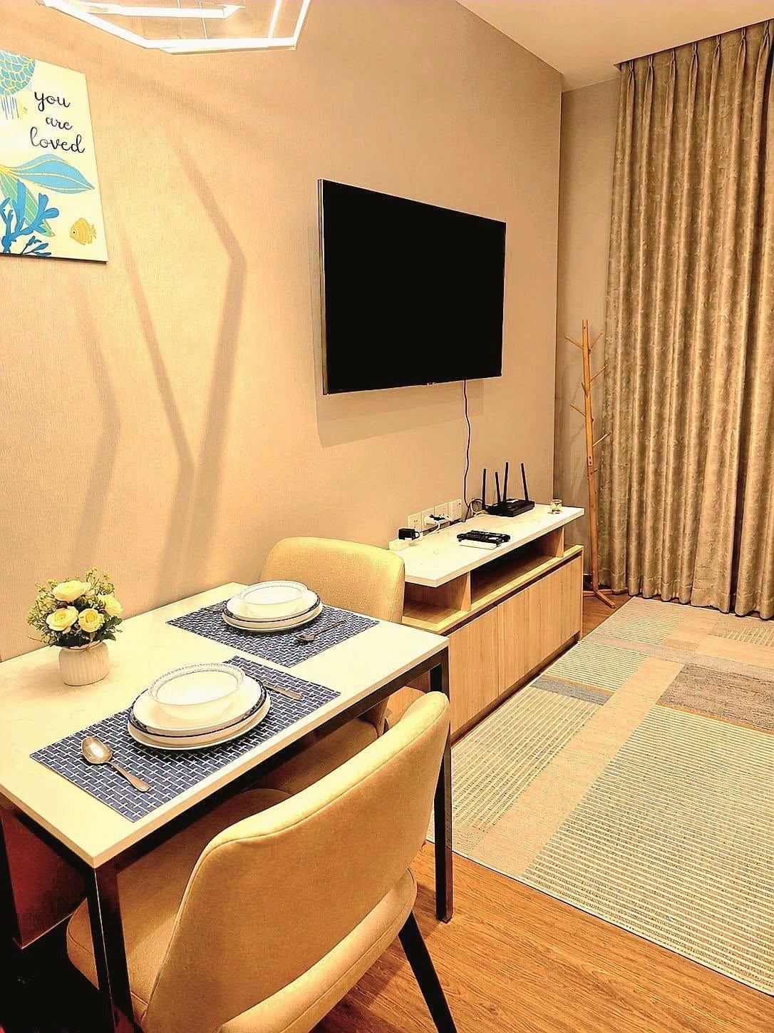 Once_Pattaya_Rent_Rdl337 Dining area with two beige upholstered chairs and a white table set with plates, a vase of white flowers, and blue placemats; wall-mounted TV nearby
