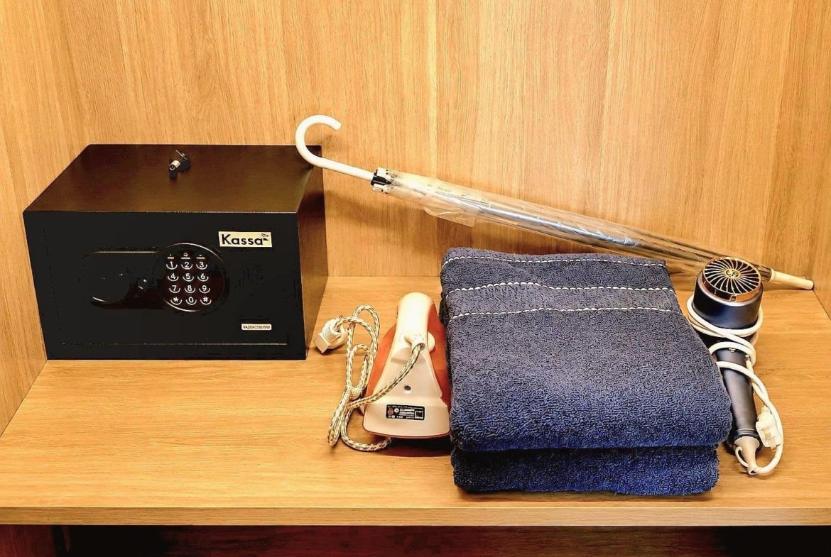 Black safe with a numeric keypad on a wooden shelf, labeled 'Kassa', beside a white curved item, an iron, and a stack of blue towels.