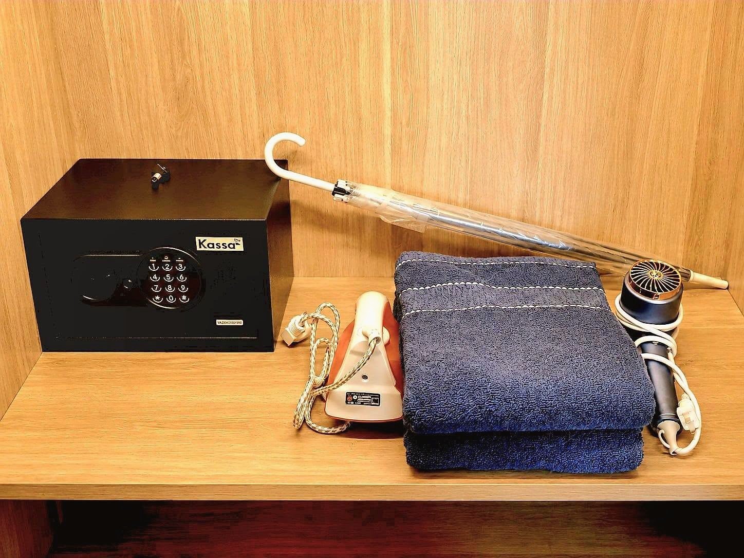 Once_Pattaya_Rent_Rdl337_10 Black safe with a numeric keypad on a wooden shelf, labeled 'Kassa', beside a white curved item, an iron, and a stack of blue towels.