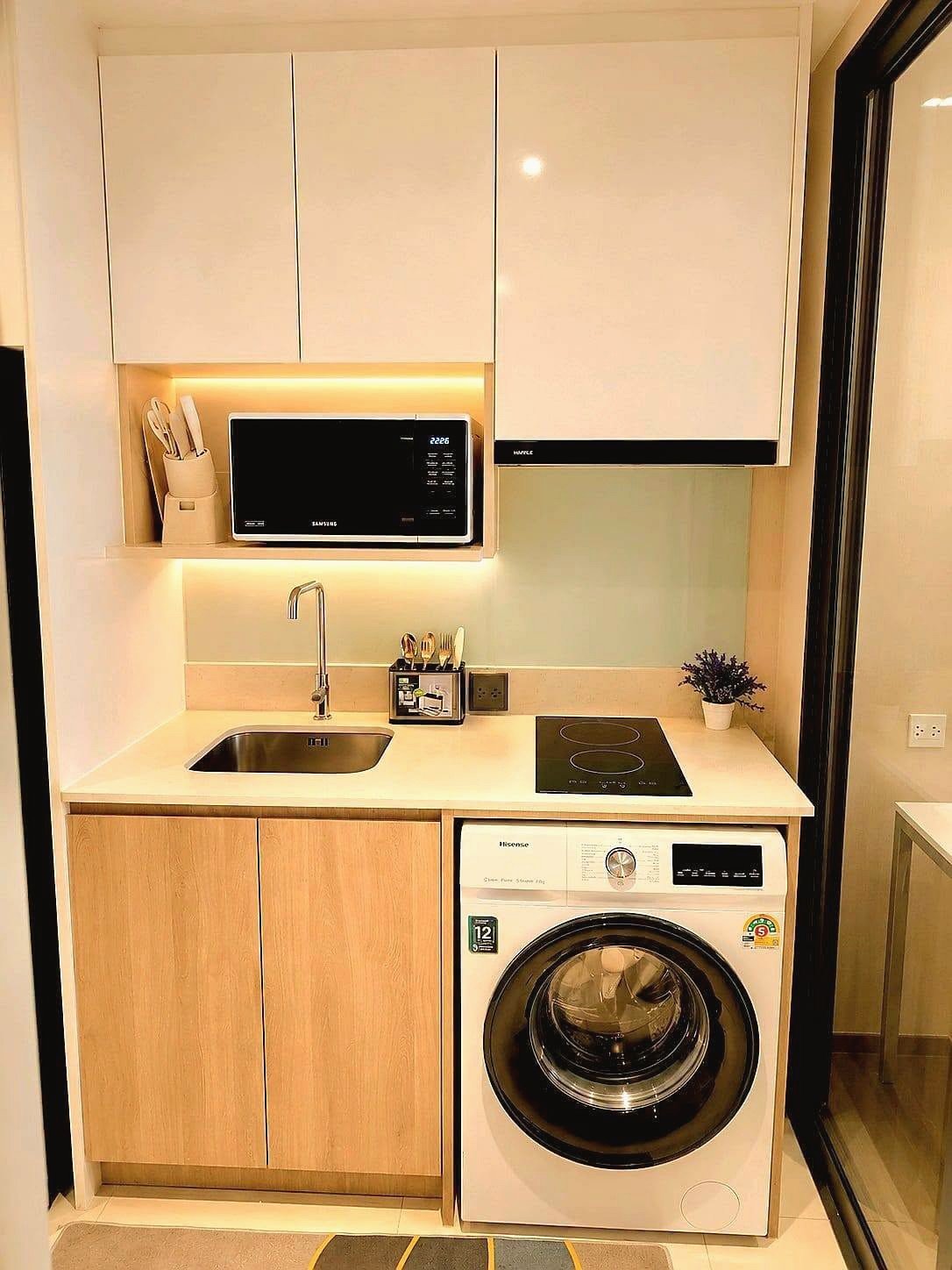 Once_Pattaya_Rent_Rdl337_2 Compact kitchen with washing machine under the counter, induction hob, sink, and white upper cabinets with under-cabinet lighting.
