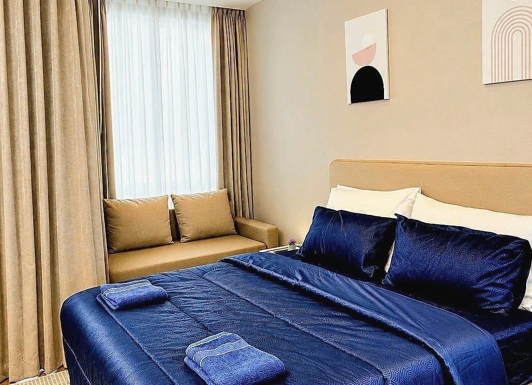 King bed with navy blue satin comforter and matching pillows in a beige-toned room with sheer and blackout curtains and two folded towels on the cover.