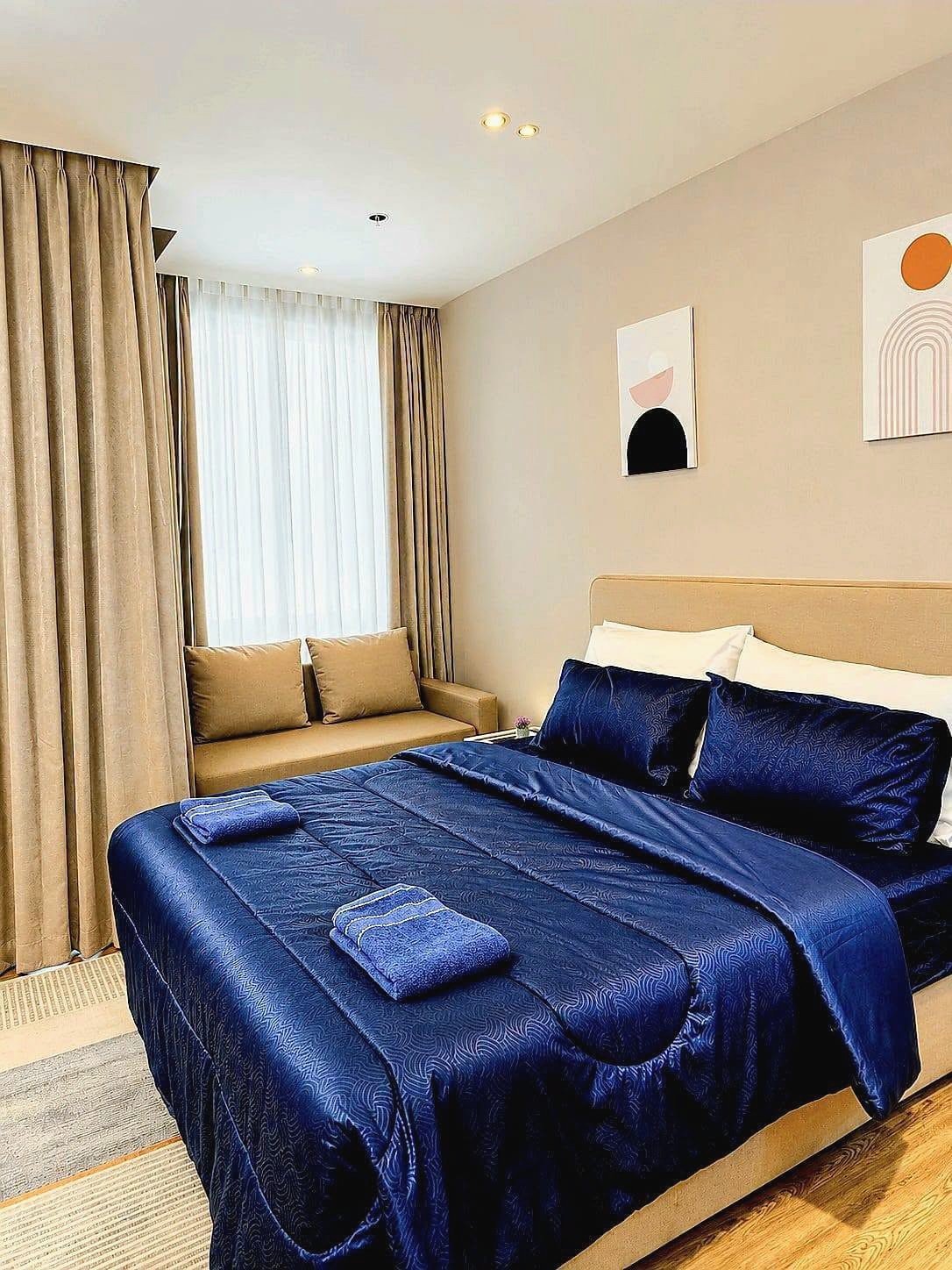 Once_Pattaya_Rent_Rdl337_3 King bed with navy blue satin comforter and matching pillows in a beige-toned room with sheer and blackout curtains and two folded towels on the cover.