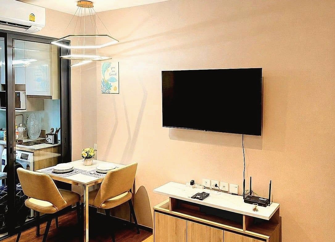 Open-plan living area with a wall-mounted TV above a wooden media console, two-beige dining chairs at a small table, and blue seating at the foreground edge; kitchen visible through glass door on the left.