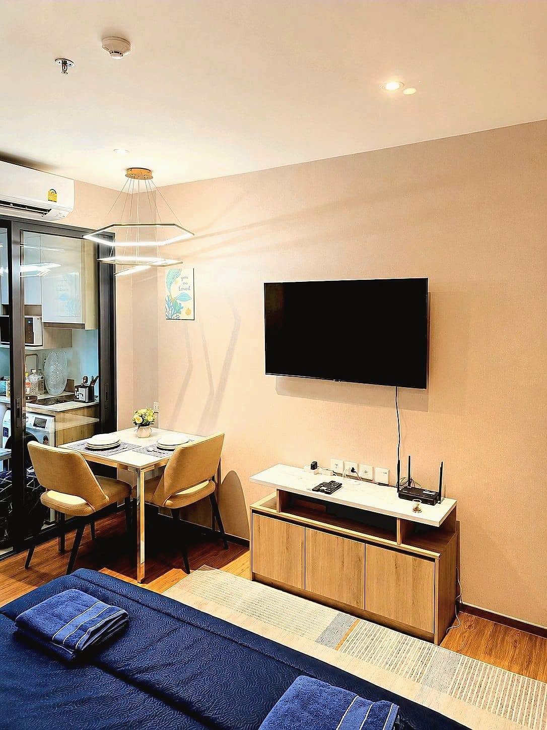 Once_Pattaya_Rent_Rdl337_4 Open-plan living area with a wall-mounted TV above a wooden media console, two-beige dining chairs at a small table, and blue seating at the foreground edge; kitchen visible through glass door on the left.