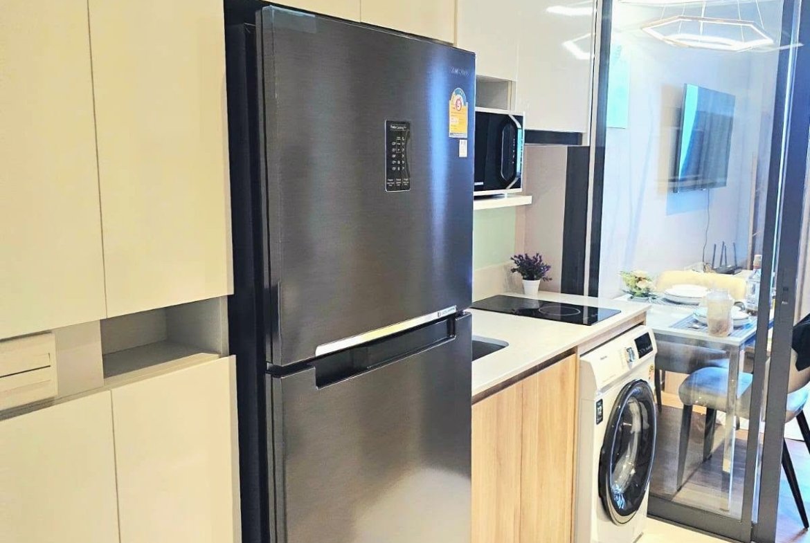 Modern kitchen with a stainless steel refrigerator, white cabinets, and a washing machine beside a glass sliding door to the living area.