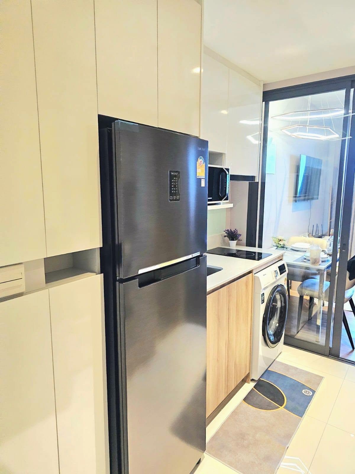 Once_Pattaya_Rent_Rdl337_5 Modern kitchen with a stainless steel refrigerator, white cabinets, and a washing machine beside a glass sliding door to the living area.
