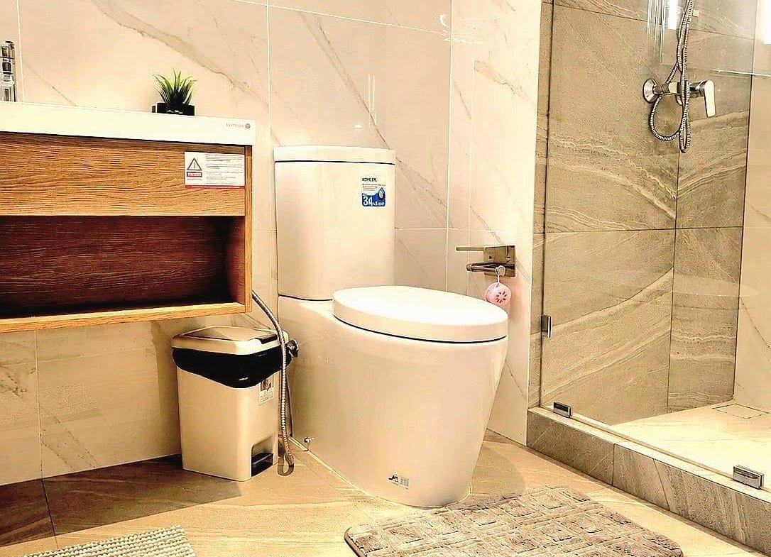 Modern bathroom with a white toilet, wooden vanity, small plant, trash bin, and a glass-enclosed shower with marble tiles.
