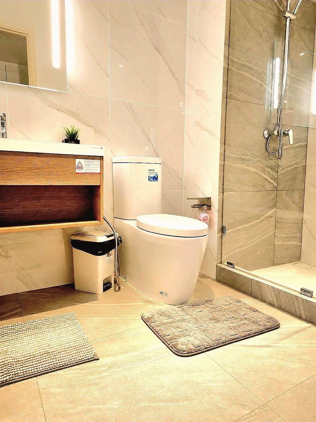 Once_Pattaya_Rent_Rdl337_9 Modern bathroom with a white toilet, wooden vanity, small plant, trash bin, and a glass-enclosed shower with marble tiles.