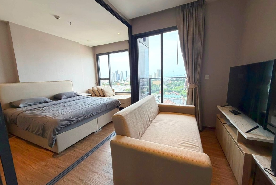 Modern hotel bedroom with two neatly made beds, a beige sofa, and a large window view of the city skyline to the right.
