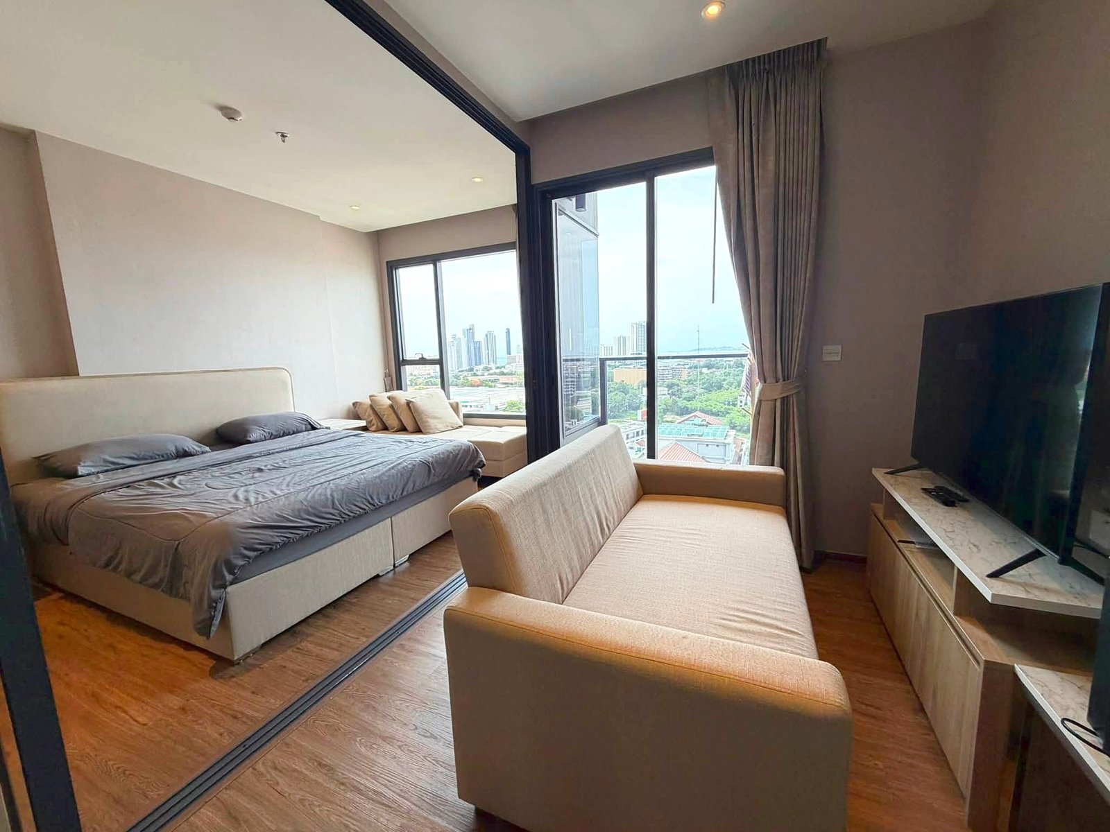 Once_Pattaya_Rent_Rdl366 Modern hotel bedroom with two neatly made beds, a beige sofa, and a large window view of the city skyline to the right.