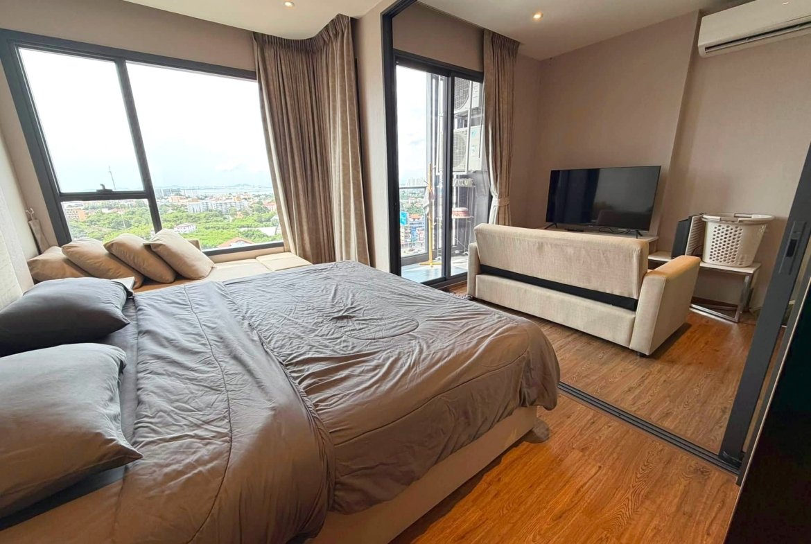 Bedroom with two full beds in brown bedding, beige sofa, and a wall-mounted TV; large windows reveal a city view in a high-rise apartment.
