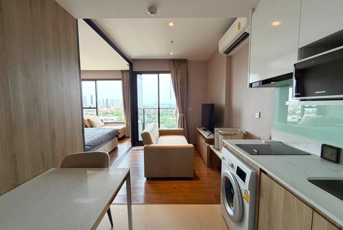 Open-plan studio apartment with kitchen on the right, living area in the center, bed alcove to the left, and large city-view windows at the back.