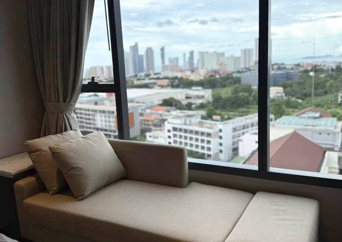 Beige window seat with cushions beside a large floor-to-ceiling window showing a city skyline outside.