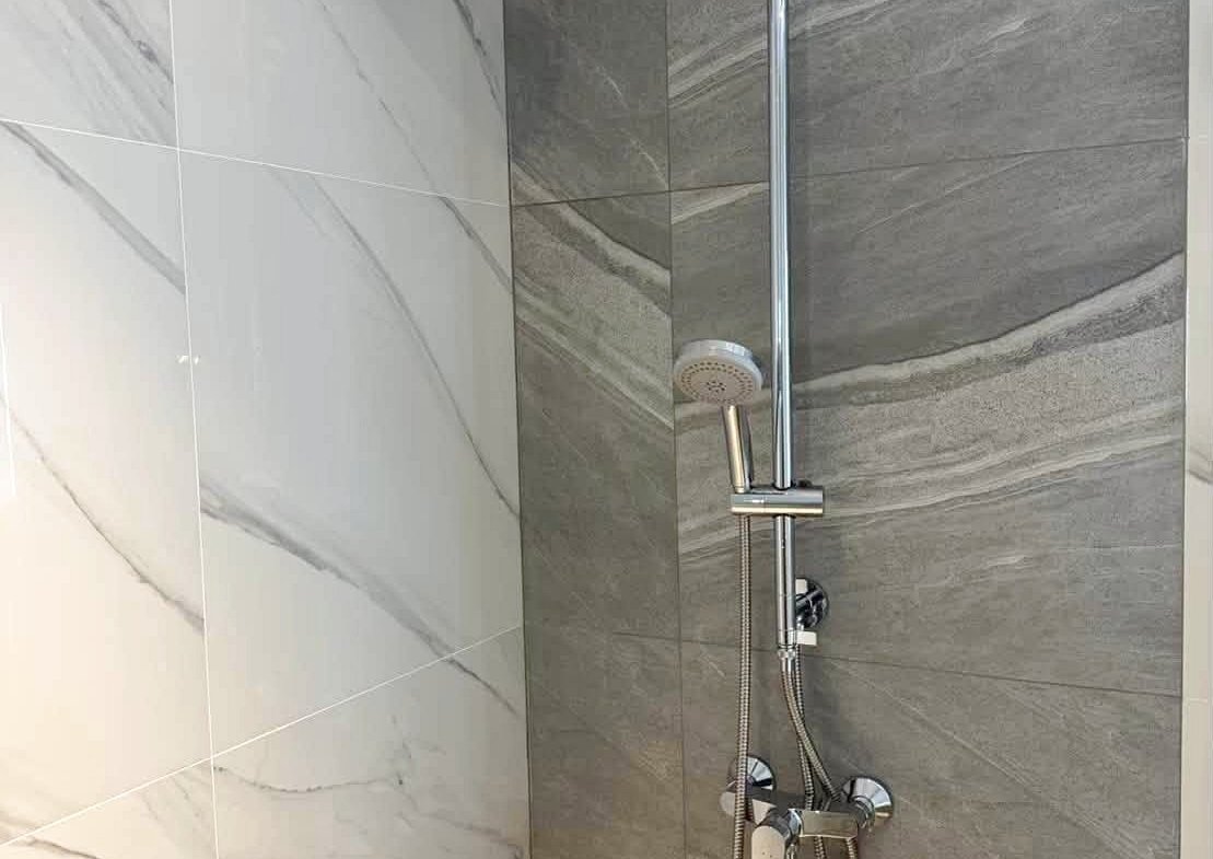 Modern shower with a curved chrome rain shower arm, a ceiling-style head, and a handheld wand on a vertical bar against gray-veined tiles.