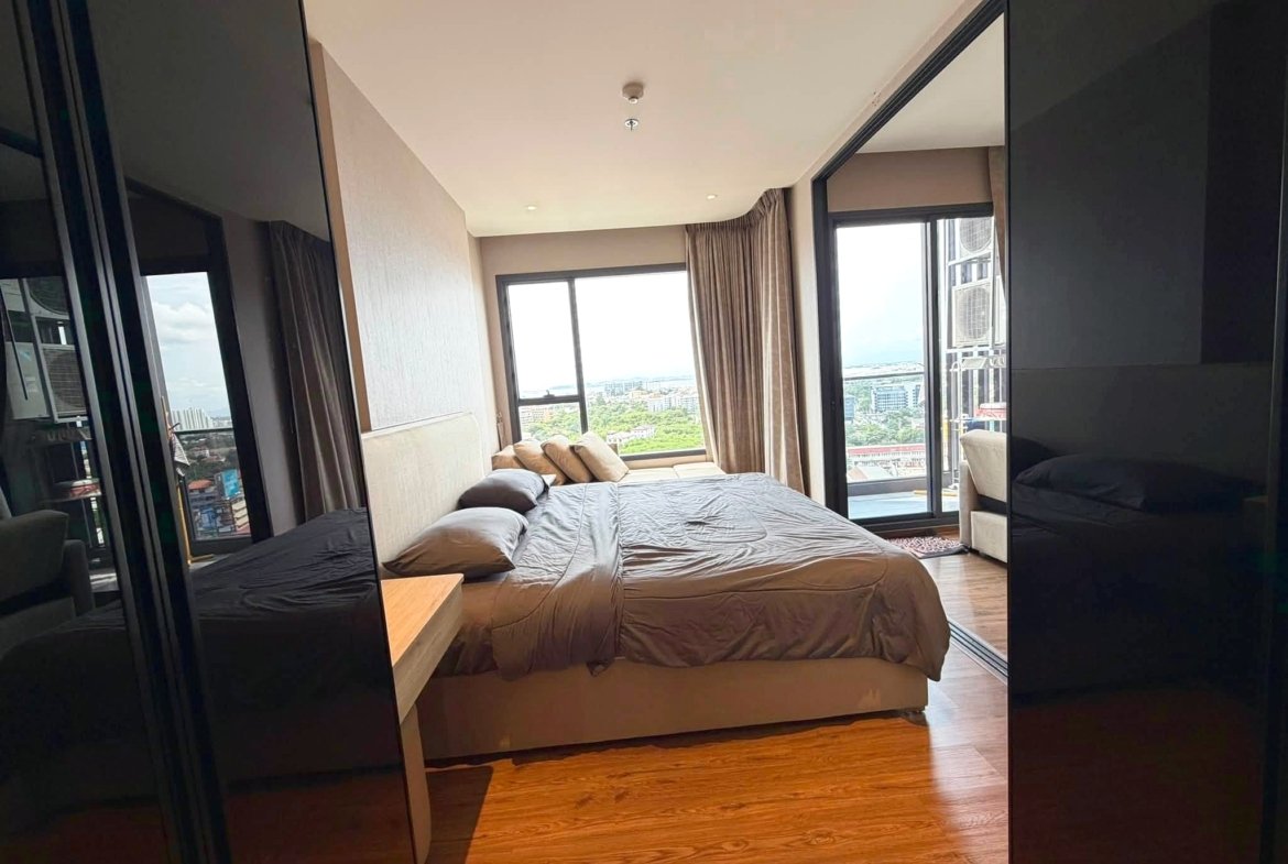 Hotel bedroom with a large bed, beige bedding, and floor-to-ceiling windows showing a city skyline.