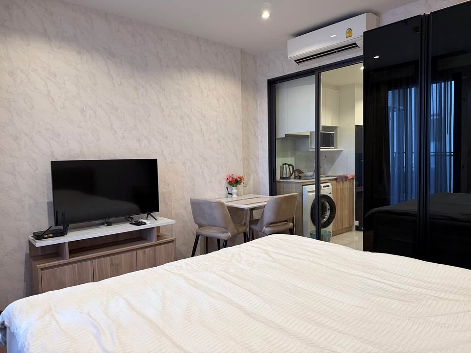 Modern hotel room: bed in foreground, flat-screen TV on a wooden stand, and a small two-person dining area beside a kitchenette through glass doors.