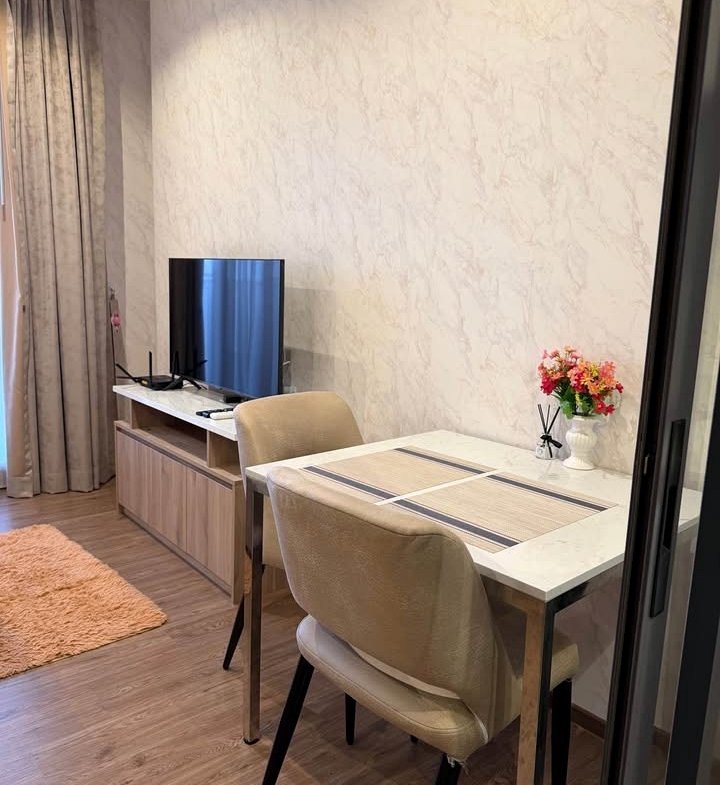 Cozy dining nook and living area with a marble-topped table for two, beige upholstered chairs, a vase of pink flowers, and a flat-screen TV on a wooden console against a pale patterned wall.