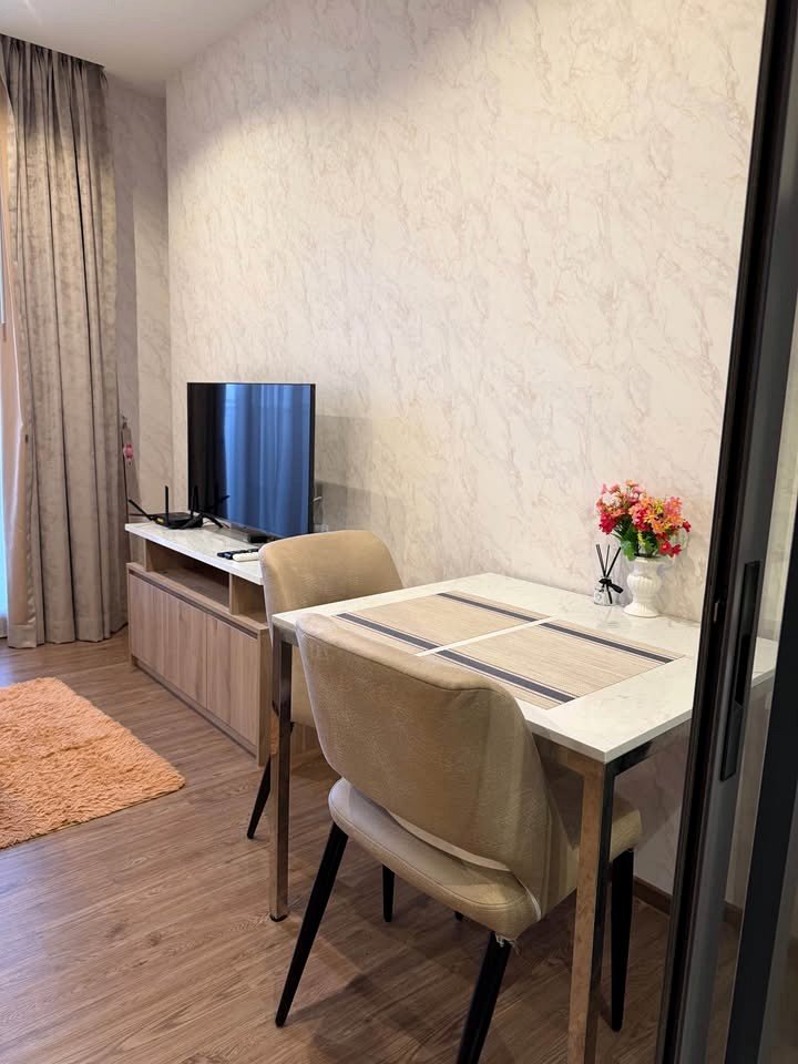 Once_Pattaya_Studio_Rdl288_2 Cozy dining nook and living area with a marble-topped table for two, beige upholstered chairs, a vase of pink flowers, and a flat-screen TV on a wooden console against a pale patterned wall.