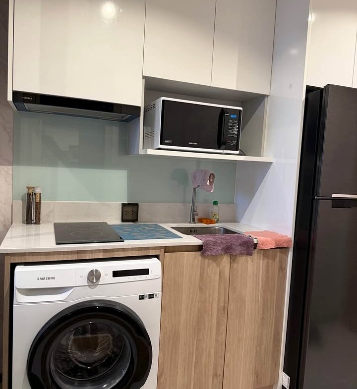 Compact kitchen with white upper cabinets, Samsung washing machine under the counter, electric stove, sink, microwave on shelf, and a tall dark fridge on the right.”