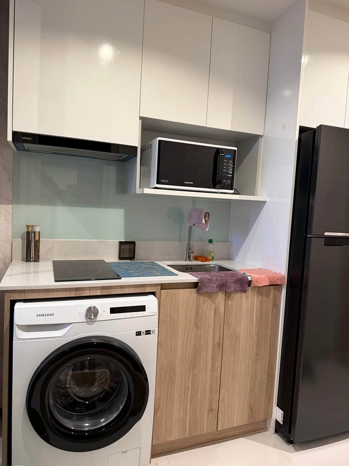 Once_Pattaya_Studio_Rdl288_3 Compact kitchen with white upper cabinets, Samsung washing machine under the counter, electric stove, sink, microwave on shelf, and a tall dark fridge on the right.”