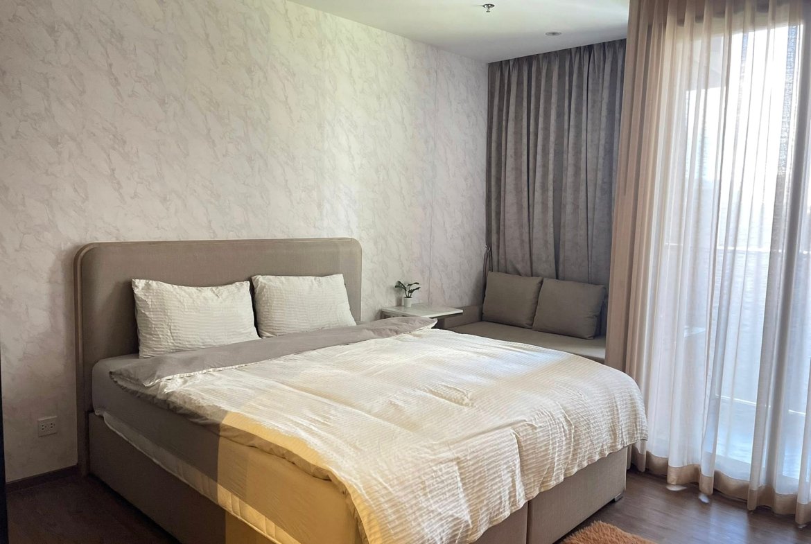 Neutral-toned bedroom with a large upholstered bed, white pillows, and textured wallpaper beside a small nightstand with a plant.