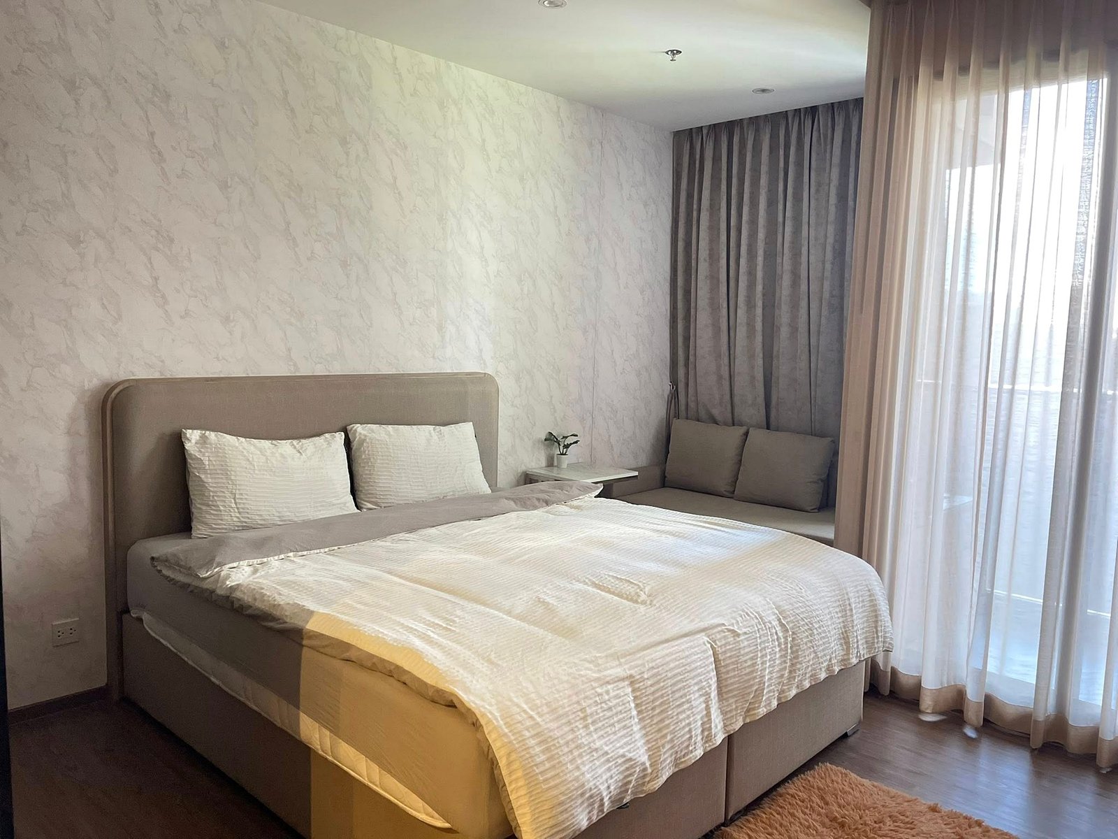 Once_Pattaya_Studio_Rdl288_4 Neutral-toned bedroom with a large upholstered bed, white pillows, and textured wallpaper beside a small nightstand with a plant.
