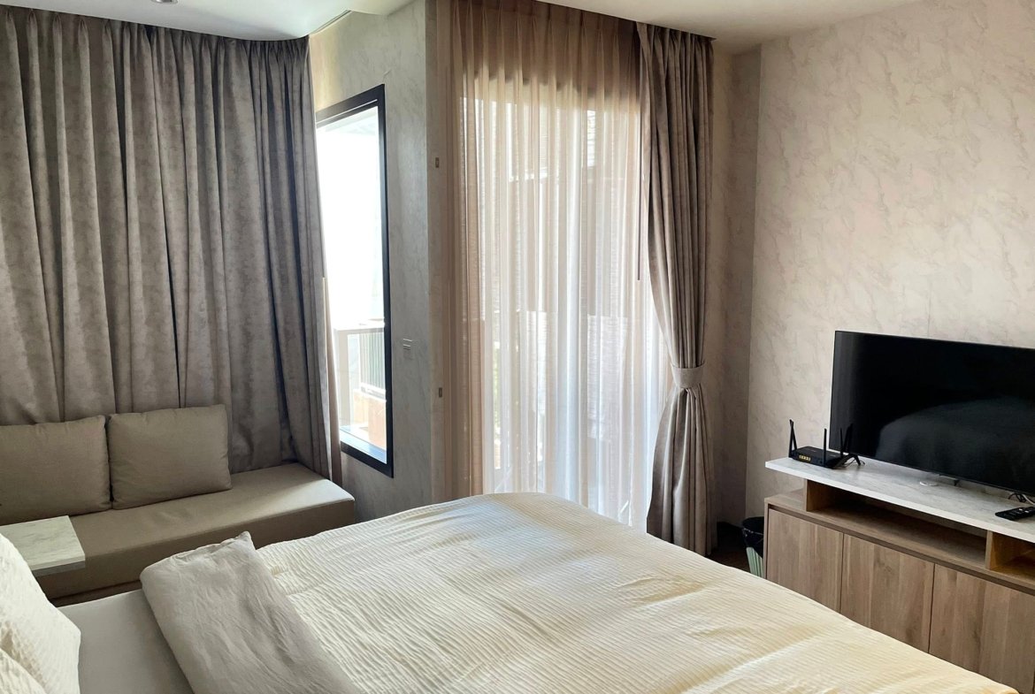 Hotel room with two neatly made twin beds and white bedding, a beige sofa along the left wall, and dark curtains over tall windows on the far wall.