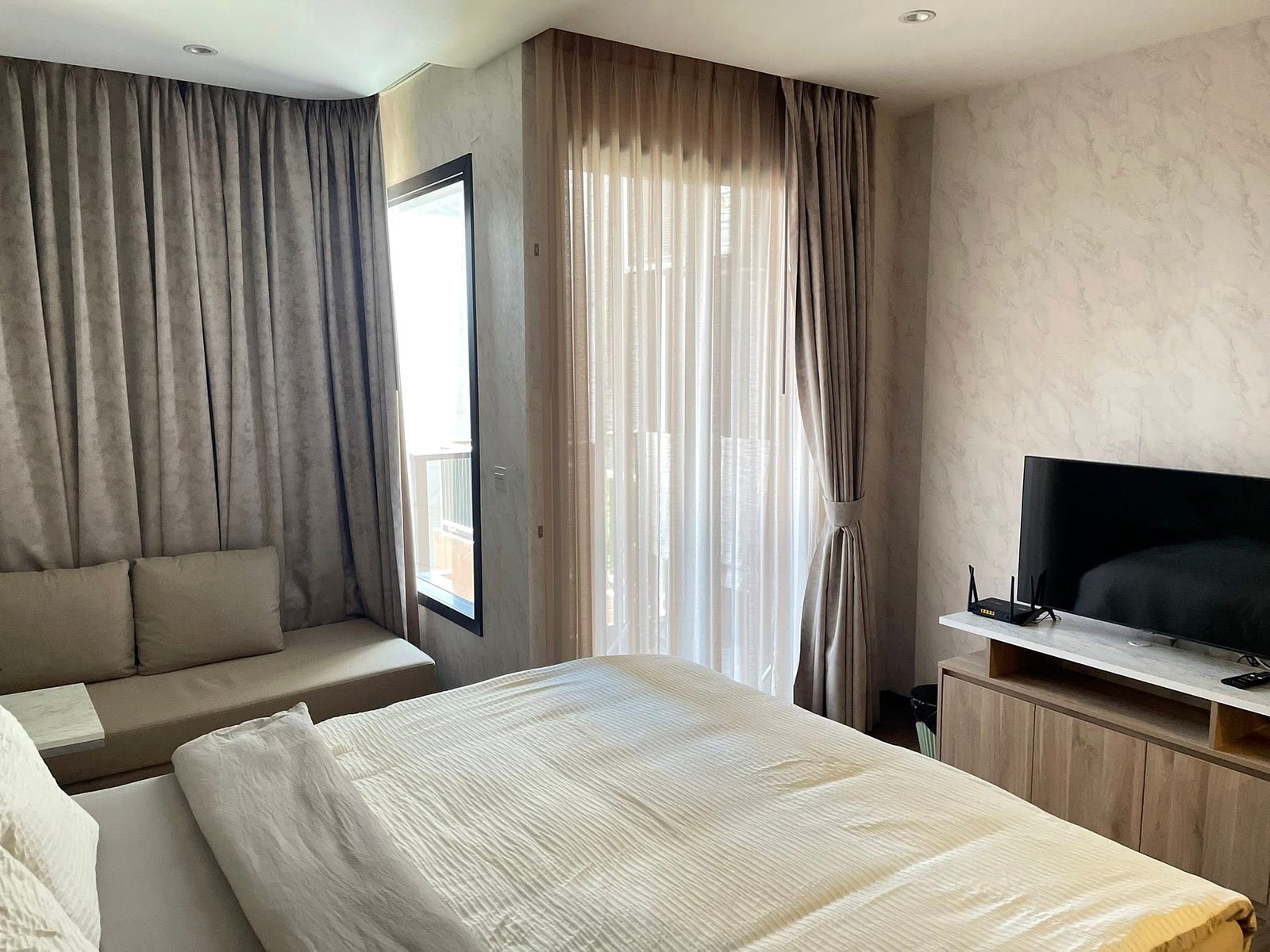 Once_Pattaya_Studio_Rdl288_5 Hotel room with two neatly made twin beds and white bedding, a beige sofa along the left wall, and dark curtains over tall windows on the far wall.