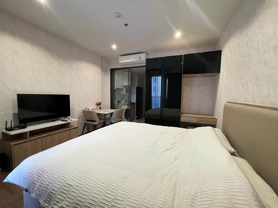Hotel room with a large white bed in the foreground, a flat-screen TV on a wooden console, and a glossy black wardrobe nearby.