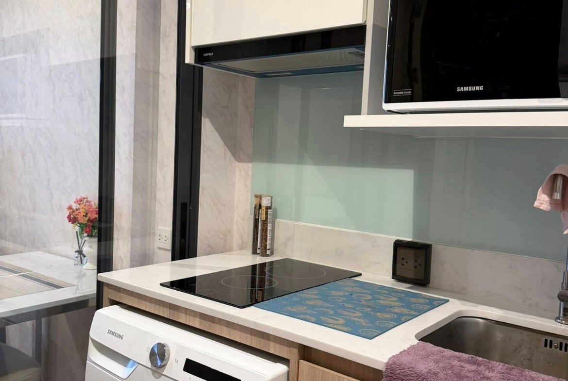 Modern kitchen corner with white cabinets, Samsung front-loading washer, black induction hob, wall-mounted microwave, glass backsplash, and a purple towel on the counter.