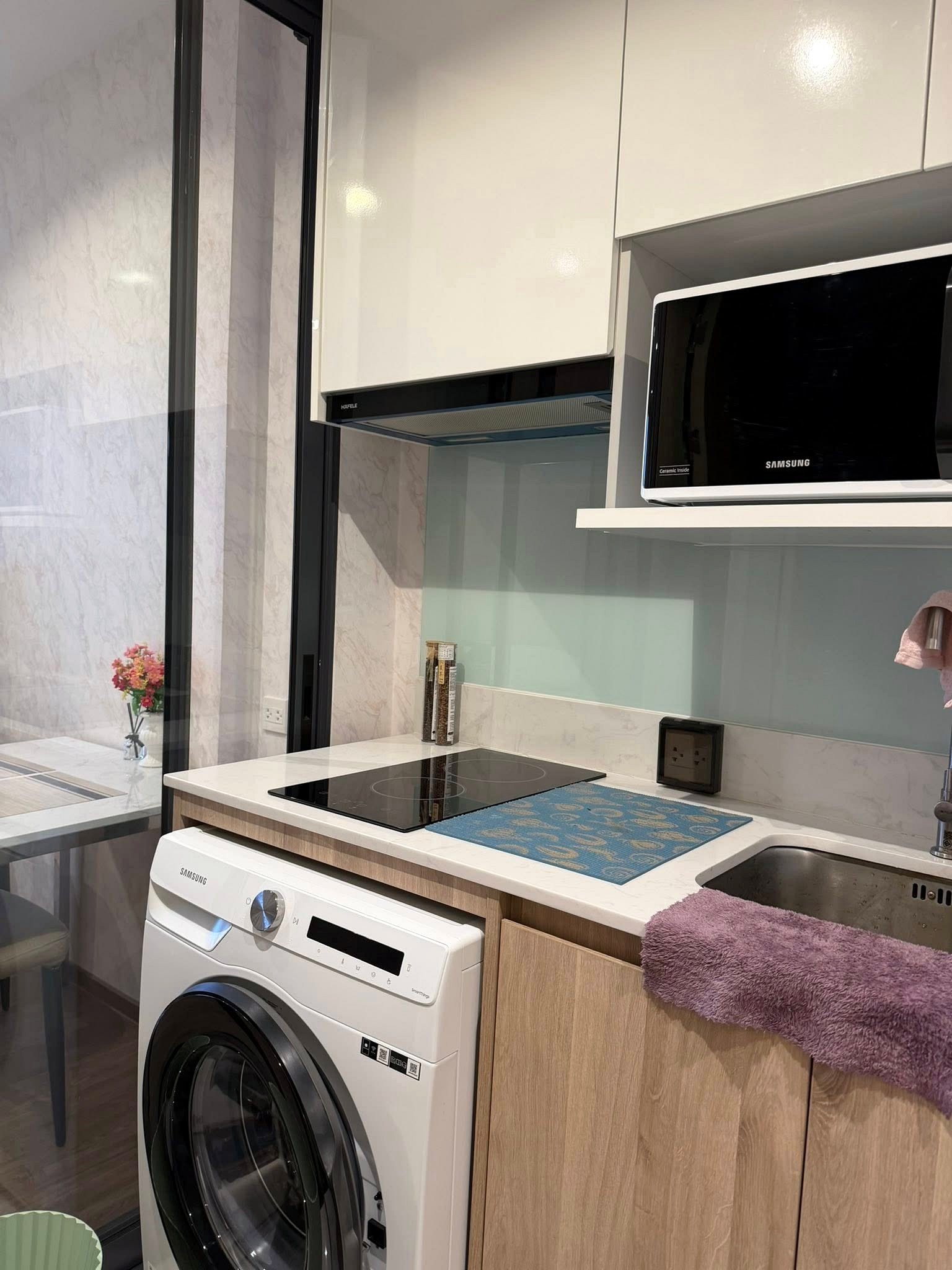 Once_Pattaya_Studio_Rdl288_8 Modern kitchen corner with white cabinets, Samsung front-loading washer, black induction hob, wall-mounted microwave, glass backsplash, and a purple towel on the counter.