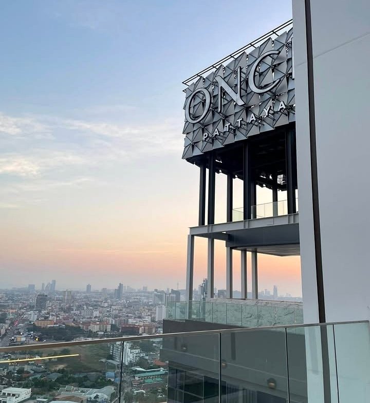 Rooftop view of a building with a large 'ONGC Pattaya' sign, overlooking a city at sunset with a glass railing in the foreground