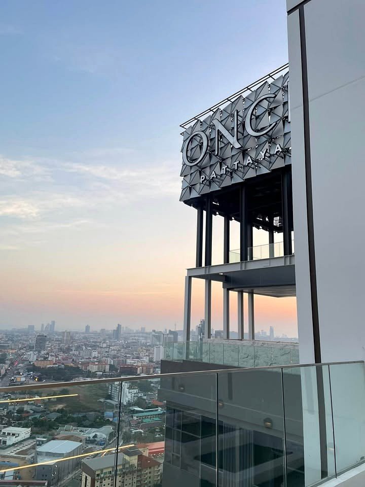 Once_Pattaya_Studio_Rdl288_9 Rooftop view of a building with a large 'ONGC Pattaya' sign, overlooking a city at sunset with a glass railing in the foreground