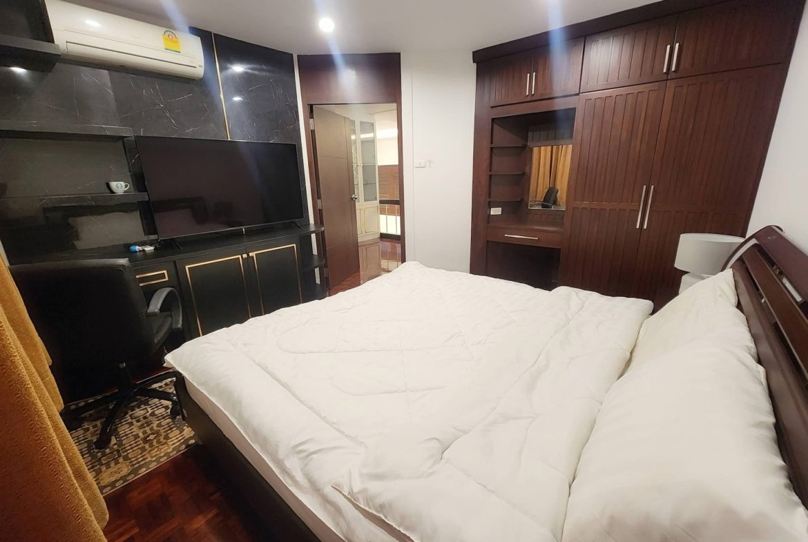 Bedroom with white quilted bed, dark wood wardrobes, and a flat-screen TV on a black marble wall.