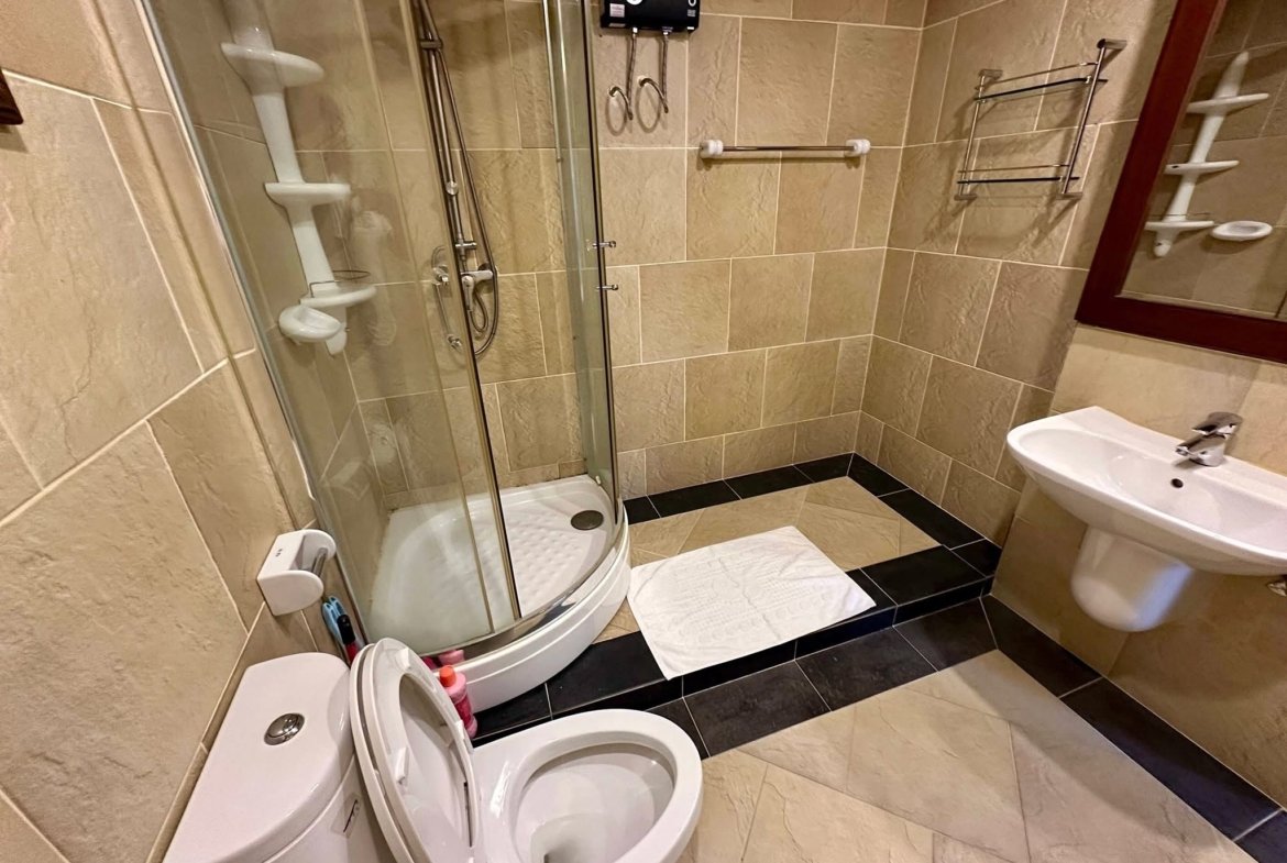 Small tiled bathroom with a curved glass shower enclosure, open toilet seat, sink, and wall mirror across beige walls and floor.