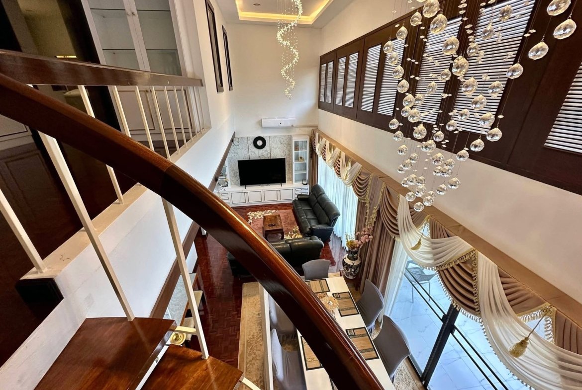 Luxurious two-story living room with a dark wood staircase, crystal chandelier, and large windows with draped curtains.