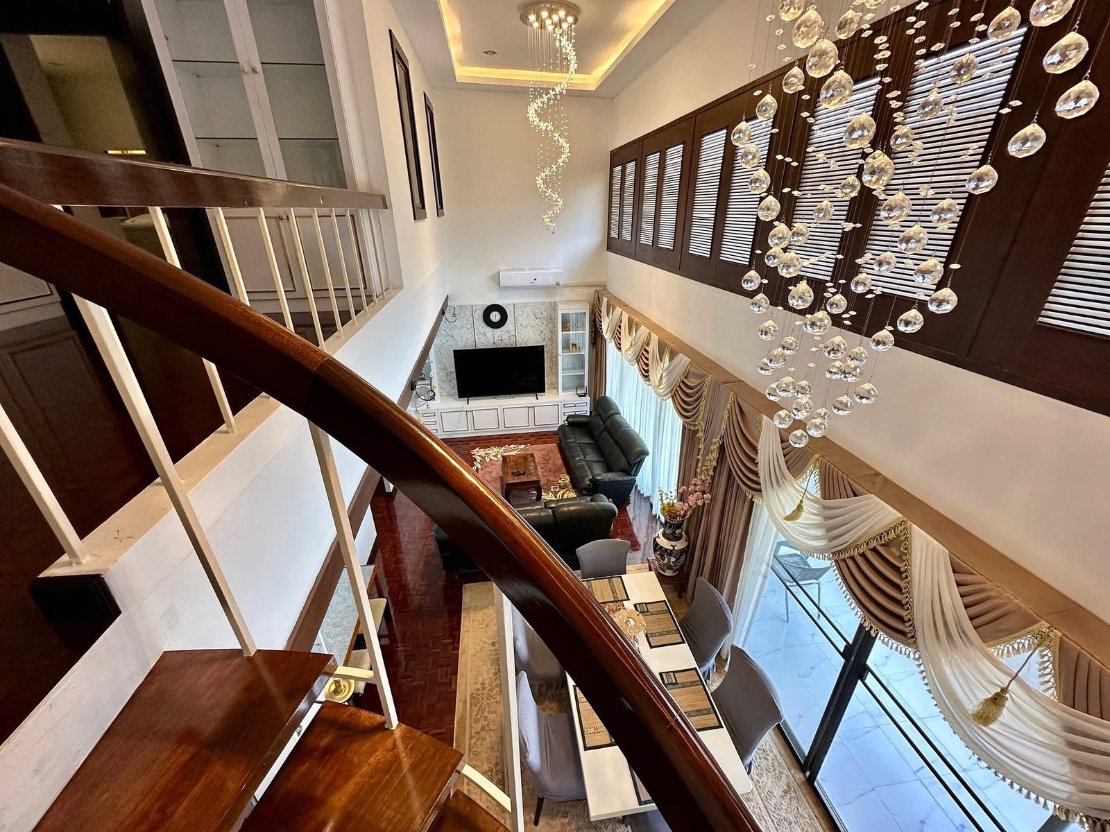 PANCHALEA_RESIDENCE_Jomtien_Rdl290_12 Luxurious two-story living room with a dark wood staircase, crystal chandelier, and large windows with draped curtains.