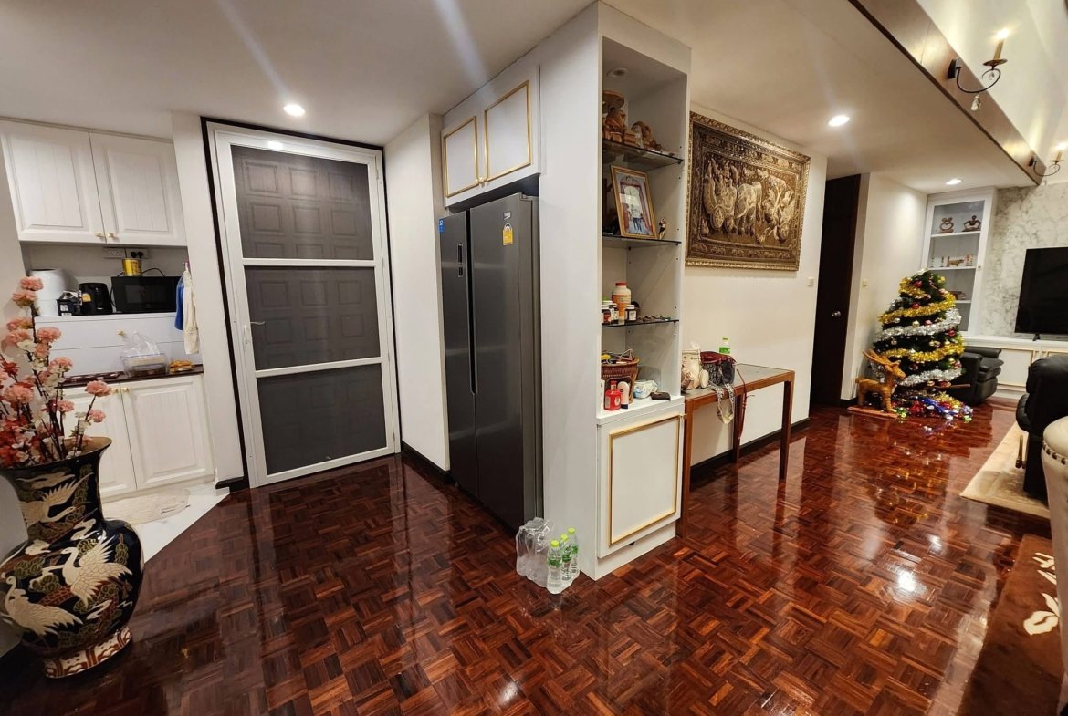 Open-plan kitchen and living area with white cabinets, a tall gray refrigerator, shelves, wall tapestry, and a decorated Christmas tree to the right.