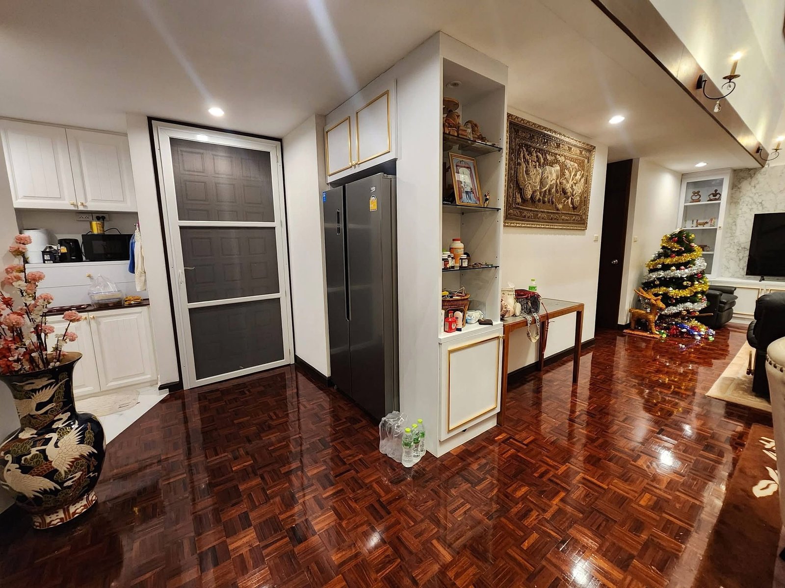 PANCHALEA_RESIDENCE_Jomtien_Rdl290_13 Open-plan kitchen and living area with white cabinets, a tall gray refrigerator, shelves, wall tapestry, and a decorated Christmas tree to the right.