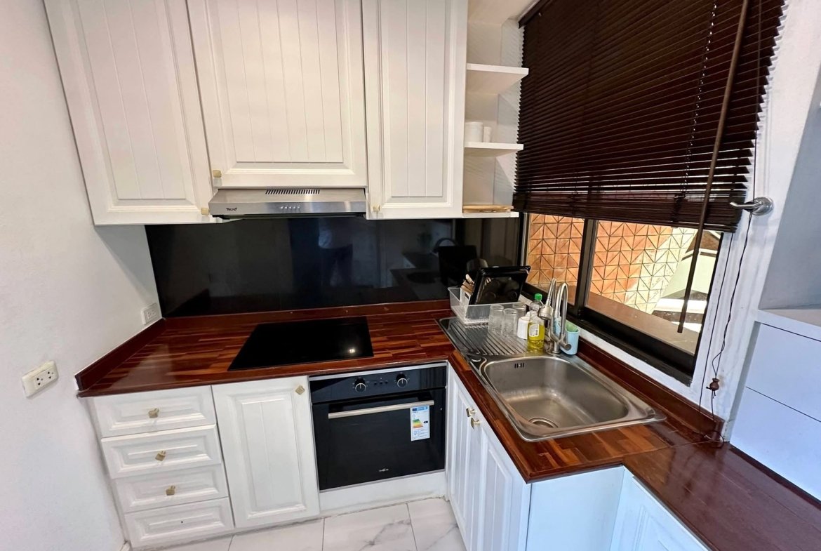 Compact kitchen with white panel cabinets, stainless steel range hood, black cooktop, built-in oven, and a sink by a window with dark blinds.