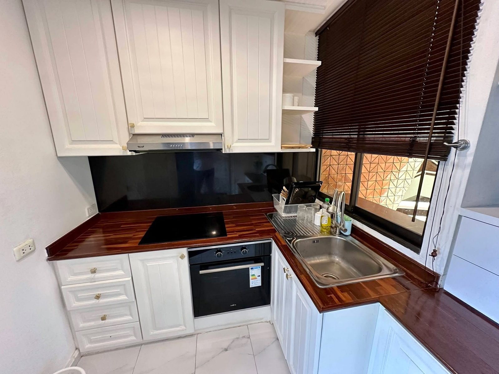 PANCHALEA_RESIDENCE_Jomtien_Rdl290_14 Compact kitchen with white panel cabinets, stainless steel range hood, black cooktop, built-in oven, and a sink by a window with dark blinds.