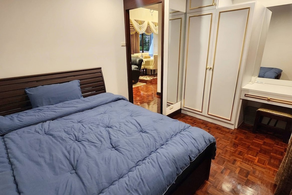 Blue quilted bed in a wood-floored bedroom with white walls and a white wardrobe with gold trim; doorway to a living area beyond.