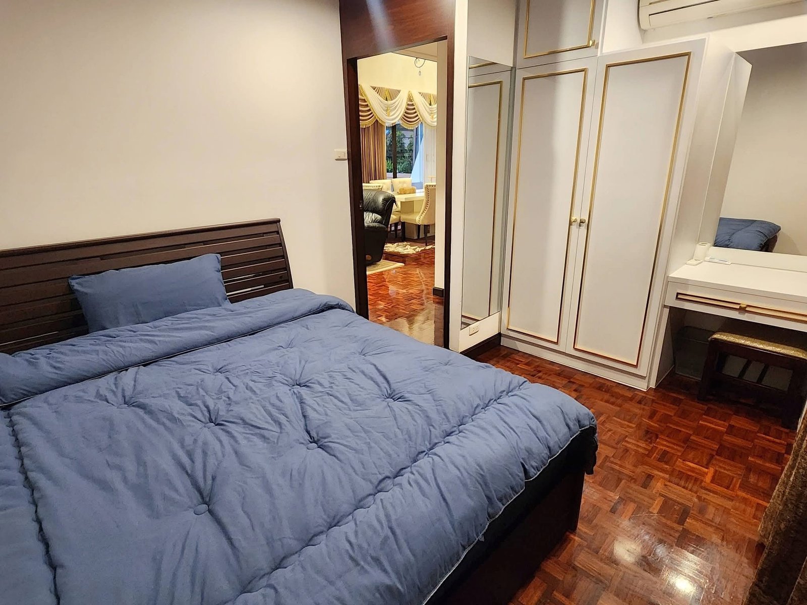 PANCHALEA_RESIDENCE_Jomtien_Rdl290_16 Blue quilted bed in a wood-floored bedroom with white walls and a white wardrobe with gold trim; doorway to a living area beyond.
