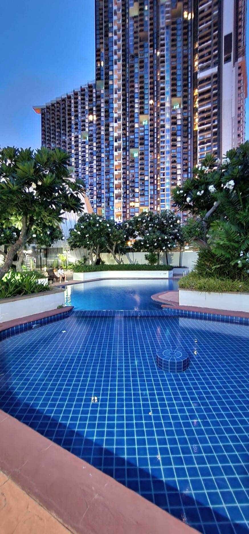 PANCHALEA_RESIDENCE_Jomtien_Rdl290_2 Curved blue-tiled pool in a courtyard with trees and a tall modern apartment building in the background.