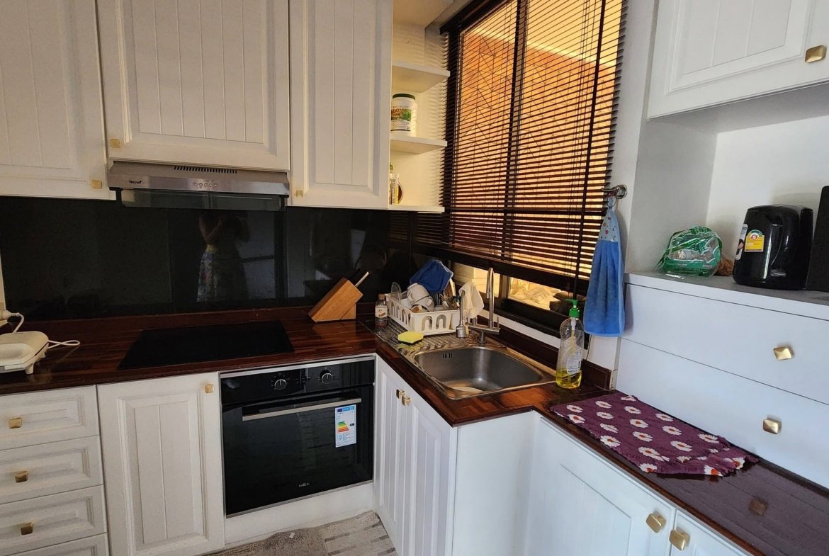 White kitchen with built-in oven, wooden countertop, dish rack by the sink, and window with brown blinds to the right.