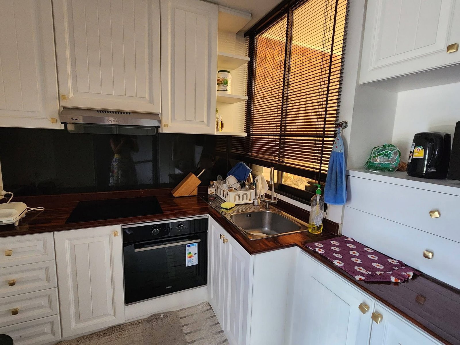 PANCHALEA_RESIDENCE_Jomtien_Rdl290_20 White kitchen with built-in oven, wooden countertop, dish rack by the sink, and window with brown blinds to the right.