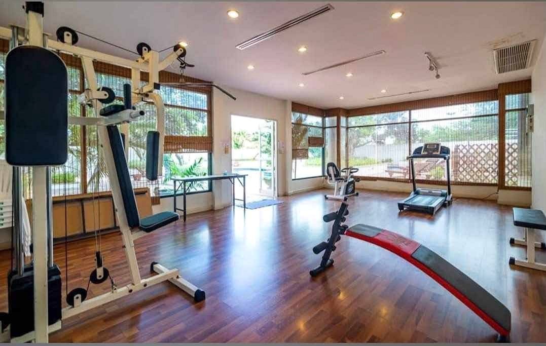 Bright gym with hardwood floor; left: multi‑station weight machine, center: curved red‑and‑black bench, back: treadmill and stationary bike by large windows.